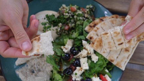 Hand breaks pita and dips it in hummus. Meze plates with salad, dips and pita. Healthy organic middle eastern cuisine. 