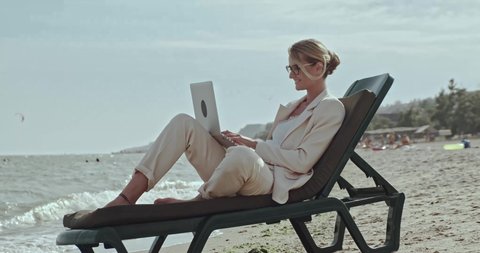 woman businessman on the beach on a sunbed in a white business suit with a laptop speaks by phone