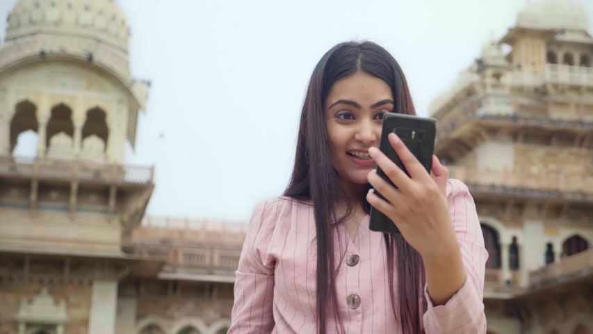 A beautiful young woman/ female is standing outdoors is holding a mobile phone in a hand and talking on an online video call. An attractive girl is communicating with a friend using a smartphone 