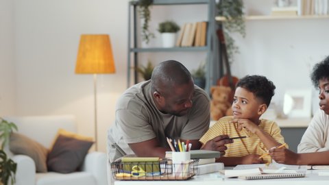 Joyous afro-american family doing homework together with little son, then looking at camera and smiling while sitting at table in the living room