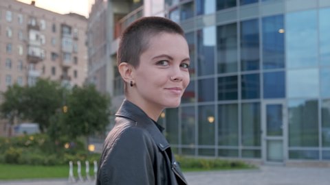 Young woman with shaved hair walking against urban background with modern buildings. Portrait of stylish woman with nose piercing at summer.