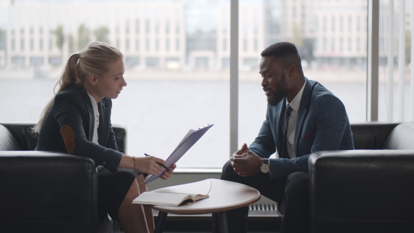 Young HR woman interviews afro-american male candidate for job sitting on couch in conference room. Sales manager holding clipboard and talking to client in modern business center Royalty-Free Stock Footage #1058132395