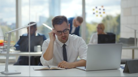 Tired young businessman feeling stressed taking off eyeglasses having eyestrain after long office work on computer. Exhausted office manager sitting at desktop with colleagues working on background