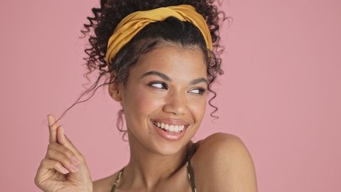 A close-up view of a smiling cheerful african american young woman is posing to the camera while touching her curly hair standing isolated over pink background