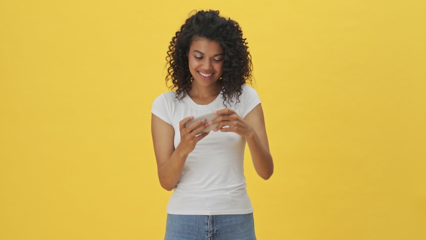 Young african emotional woman playing games by mobile phone isolated over yellow wall background