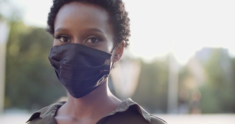 Portrait of isolated ethnic girl standing in street putting on black medical mask on female African face, protecting herself from covid virus, pandemic time, pollen allergy seasonal disease, close up