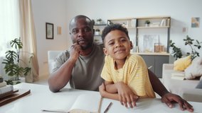 Joyous African American father and cute little son smiling, waving and talking at camera while chatting on online video call at home during isolation