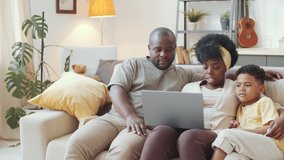 Tracking shot of happy afro-american family sitting together on couch in living room and watching movie online on laptop while staying together at home - Powered by Shutterstock - Get 15% off with code: PIKWIZARD15