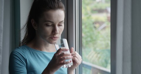 Woman Taking a Pill and Drinking a Glass of Water Standing by the Window