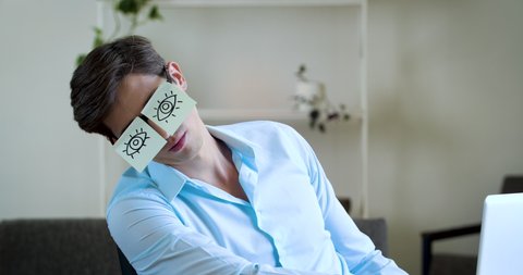 Close up sleepy funny office manager sitting workplace asleep with stickers on eyes. Portrait comic humorous lazy businessman student napping on desk in office, wears glasses with paper sticky notes
