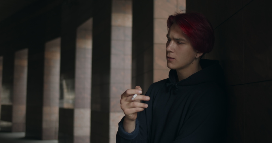 Crop view of millennial guy in depression smoking cigarette and thinking. Young hipster person with red dyed hair and earring looking aside while standing at urban street