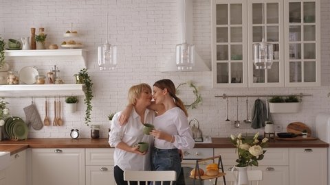 A happy elderly mother embraces her daughter in the bright kitchen, they hold green mugs of tea in their hands