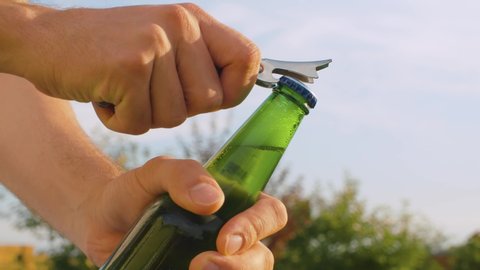 Close up male hand opening bottle of beer using opener outdoors on sky background. The camera slowly moving on the slider