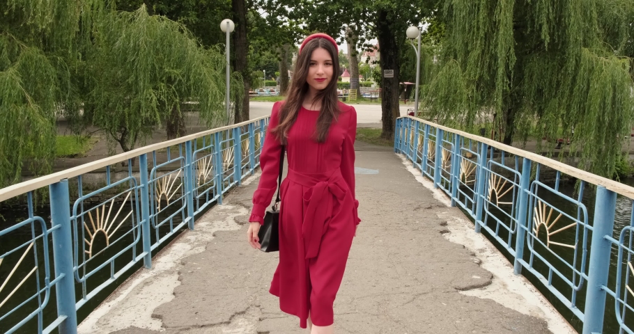 Gorgeous brunette in red touches her hair and crossing bridge in the park