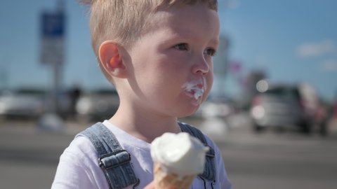 Portrait of blond caucasian boy in white t-shirt and denim overalls, eating and enjoying creamy cold ice cream in waffle cone cup, sitting outside in sunny weather.