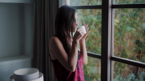 Side view of woman in pajamas drinking coffee in morning and looking out window, slow motion. Greenery on background. Girl relaxing and enjoying