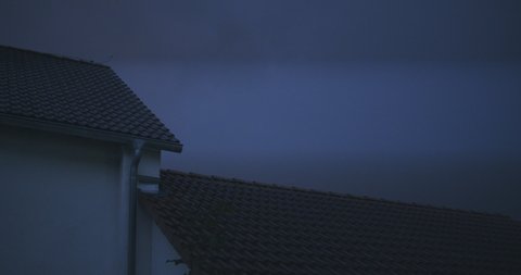 Medium white shot of a roof in an suburban neighborhood during a thunderstorm. Night sky gets bright due to lightning.