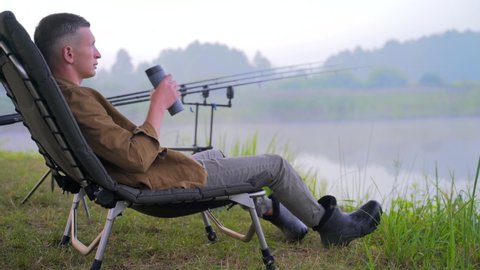 young stylish fisherman guy sitting on a fishing chair and drinking a drink, tea coffee from a thermos. background of lake fishing rods and trees nature. Hipster man on vacation. Active recreation 