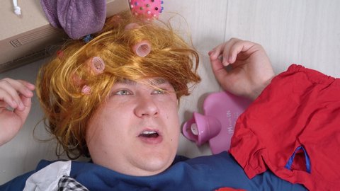 Crazy guy in shabby wig with pink curlers is lying on floor with scattered stuff and heating pad. Attempts to put things in order, storage in small apartment. Psychological problems with gathering