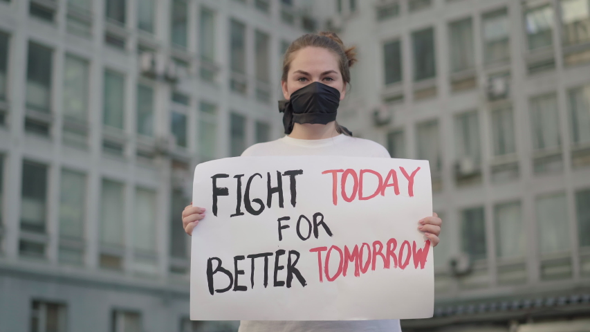 Beautiful woman in face mask standing in urban city with banner and looking at camera. Portrait of female Caucasian demonstrator protesting against social inequality and rights violation. Royalty-Free Stock Footage #1058172961