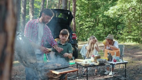 Family adventures in the woods. Picnic party. Father and son preparing grilled burger cutlets on braizer. Beautiful similar mother and daughter cooking vegetables at table.