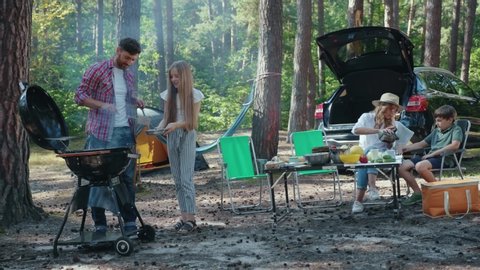 Caucasian family resting together having barbecue party in beautiful wood landscape. Affectionate family members enjoying fresh grilled food on picnic.