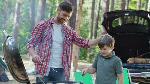 Cheerful boy helps his bearded dad cooking and roasting beef in brazier griller outdoors. Family arrive in forest for summer camping. Together. Picnic.