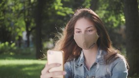 lady on video call outdoor in the park during social distancing pandemic of coronavirus wearing protective face mask using smart phone and video conferencing technology.