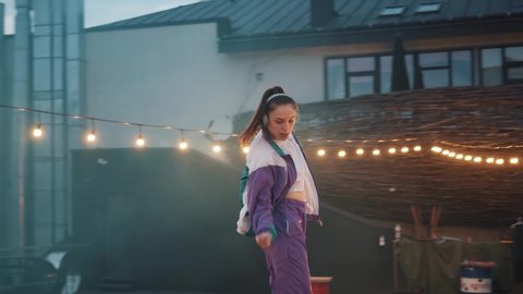 Young excited woman in sports wear with earphones dancing outside the roof with lights glare on blur background. Freestyle dance improvisation. Brunette moving by music rhythm. 4K