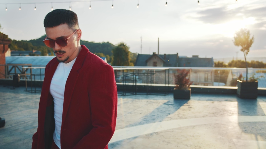 Attractive brunette fashion man in sunglasses and red cherry suit dancing on roof terrace outdoor on sunny background. Professional male dancer performing freestyle dance show. Sunlight rising. 4K