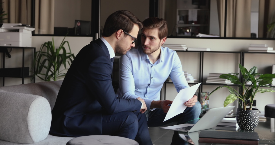 Young smiling male employee holding paper document, discussing contract with confident partner in formal wear, sitting on couch in modern office. Happy businessmen shaking hands, making agreement. Royalty-Free Stock Footage #1058203663
