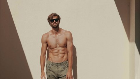 Handsome smiling strong man. Healthy athletic fitness model posing near white wall in jeans. Sexy fashion male with naked nude torso. Lambersexual outdoors.
He straightens his hair in sunglasses