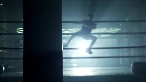 Kickboxer or Muay thai fighter training in low light gym . Silhouette on dark background . Boxer training in ring . Cinematic slow motion shot of a professional young man practicing in boxing ring