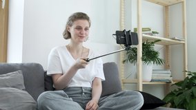 caucasian girl sits on couch, holds selfie stick with smartphone and talks to her family. female having video chat with relatives using smartphone. Modern technology, communication