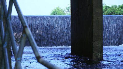 Camera moving to left. Shade and view through channel of watercourse under the bridge. Old concrete dam with water flowing rapids. During the rainy season in rural Thailand.