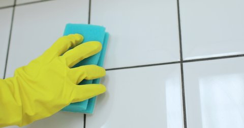 Unrecognizable maid is cleaning wall. Hand in yellow glove is washing white tile using blue sponge, closeup view. Concept of cleaning, housekeeping, housecleaning. Professional cleaning of home.