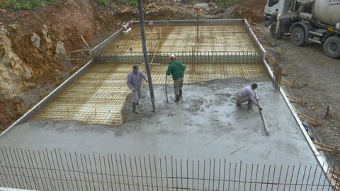 LJUBLJANA, SLOVENIA, APRIL 2020: A crew of contractors pours wet concrete over the metal wiring placed on the ground floor of a house under construction. Group of builders pours a concrete slab.