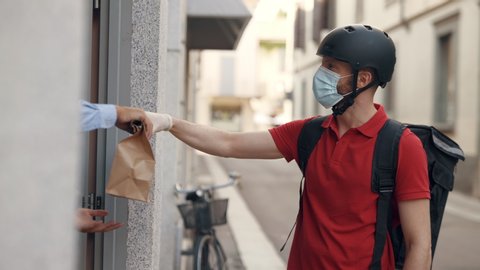 An young courier with a protective mask and gloves is delivering a grocery order with fresh food to a customer home with safety. Concept of courier, home delivery, e-commerce, online shopping, covid