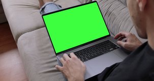 Man sitting on sofa in front of MacBook Pro laptop with green screen, chroma key, makes video call to friends, family, working on keyboard