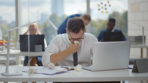 Unhealthy male employee sneezing while working in workplace in office. Sick young businessman working on computer using napkins. Tired ill managers sitting at desktop