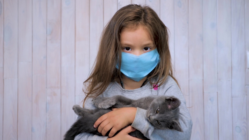 Stay at home in quarantine, funny kid with kitten at home during COVID-19 coronavirus pandemic. Little girl in medical mask for protection to corona virus indoors looking at camera on wood grey wall - Powered by Shutterstock - Get 15% off with code: PIKWIZARD15