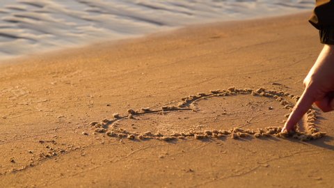 Close up view of woman hand drawing heart with finger on the sand at the beach. Romantic love symbol at tropical seashore at sunset
