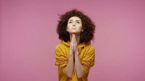 Please god, I'm begging! Upset young woman afro hairstyle in hoodie appealing to heaven with pleading imploring eyes, keeping prayer gesture and asking help. indoor  isolated on pink background