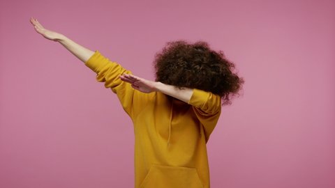 Trendy young woman afro hairstyle in hoodie performing dab dance with serious calm face and making dabbing movement with hands up, internet meme gesture. indoor studio shot isolated on pink background