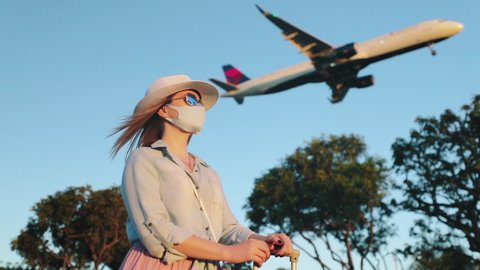 Slow motion woman in face mask is enjoying the sunset with landing airplane on background. Safe travels under COVID-19. Aircraft is arriving at the airport during coronavirus pandemic on sunny evening