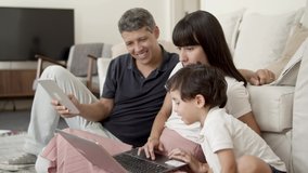 Focused parents couple and little son sitting together on apartment floor, using tablet and laptop pc. Family and internet communication concept
