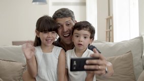 Cheerful dad and two cute kids waving hello at webcam, smiling and speaking while sitting on couch all together. Father holding phone. Medium shot. Video call concept