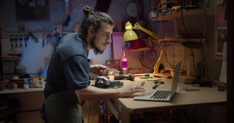 Young handsome luthier beginner searching online using computer how to use instrument, Hispanic male joiner working with wooden planks creating musical guitar instrument at studio in evening