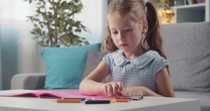 Happy little kid in blue dress drawing with colorful pencils in album. Smiling girl with two ponytails developing creativity during free time at home.