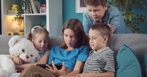 Happy children in casual clothing relaxing together on grey couch and playing games on digital tablet. Four little friends enjoying time at home with modern gadget.